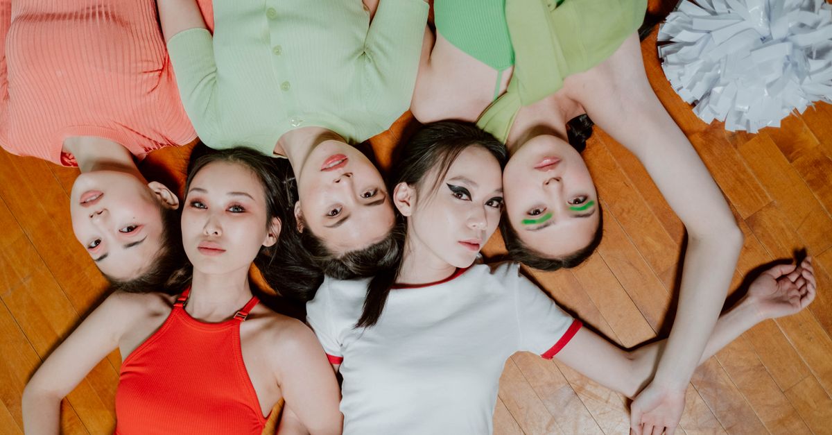 Five women in vibrant tops lying together on a wooden floor, viewed from above.